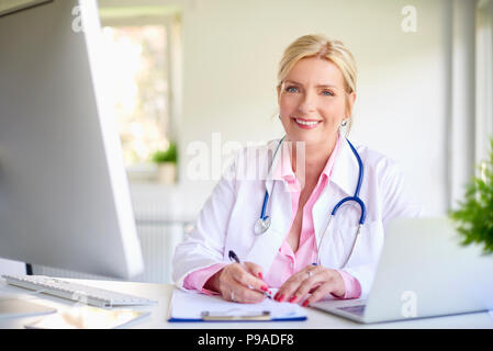 Close-up verticale di senior medico donna seduta all'ufficio del medico al banco nella parte anteriore del computer portatile e fare alcune pratiche. Foto Stock