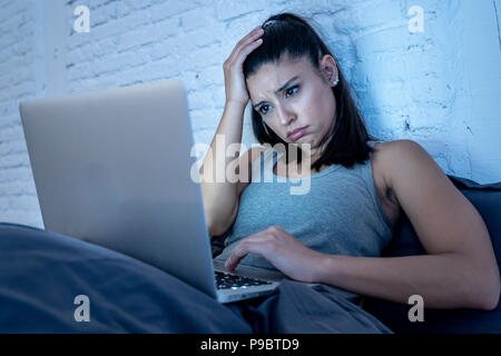 Bellissimo il latino internet addicted donna che lavorano e navigare sul suo computer annoiato, insonne e stanco nel suo letto a casa a tarda notte in camera oscura l Foto Stock