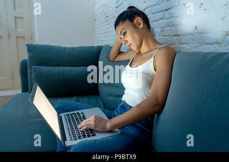 Giovani belle ispanico internet addict donna in abiti casual seduti sul divano a lavorare annoiato e stanco con computer portatile a tarda notte in da Foto Stock