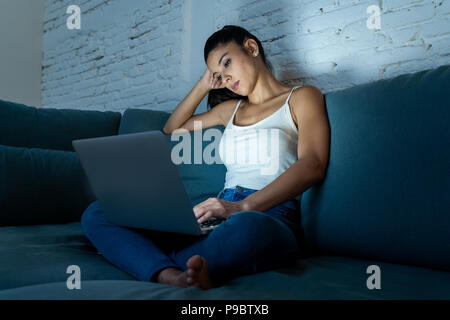 Giovani belle ispanico internet addict donna in abiti casual seduti sul divano a lavorare annoiato e stanco con computer portatile a tarda notte in da Foto Stock