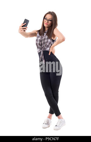 Carino giovane bella signora elegante con gli occhiali tenendo selfie foto guardando al telefono cellulare. Corpo pieno isolati su sfondo bianco. Foto Stock