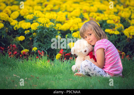 Adorabile ragazza toddler abbracciando il suo orsetto preferito in estate parco sulla bella giornata di sole con fiori gialli in background Foto Stock