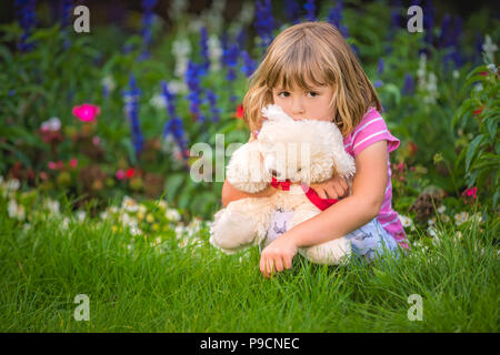 Adorabile ragazza toddler abbracciando il suo orsetto preferito in estate parco sulla bella giornata di sole con fiori gialli in background Foto Stock