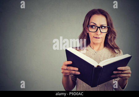 Giovane donna curiosa in bicchieri guardando lontano tenendo premuto e la lettura di libro con storie segrete su sfondo grigio Foto Stock
