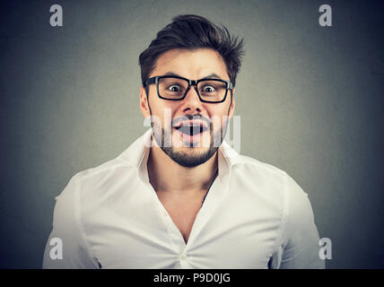 Adulto Uomo in camicia bianca e occhiali cerca storditi in telecamera con la bocca aperta su uno sfondo grigio Foto Stock