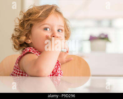 Bellissimo bambino biondo con gli occhi blu sorridente che copre la bocca a casa. Foto Stock