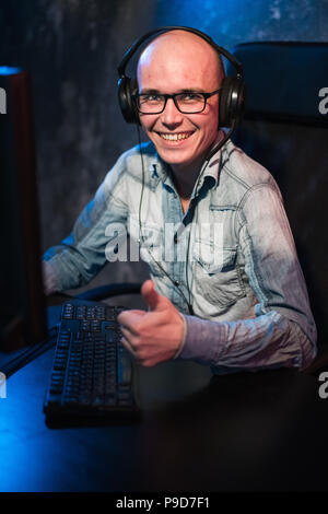 Ritratto di un giovane gioiosa gamer per giocare con i videogiochi in casa su un computer. Gamer in cuffie è felice e che mostra i pollici fino gesto perché ha vinto il gioco. Giochi di Computer concetto. Foto Stock