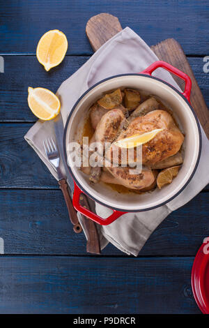 Il pollo al forno con limone e rosmarino in un rosso in ghisa. In legno di colore blu e sfondo grigio asciugamano. Coltello e forchetta. Spazio libero per il testo Foto Stock