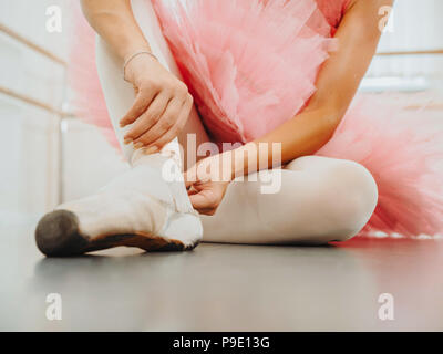 Giovane ballerina in tutù rosa avvolge il costume di nastri di seta bianca di soft top scarpe da ballo pointe e li collega. La donna la preparazione per la danza lezioni di training in palestra. Foto Stock