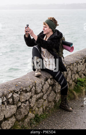 Giovane donna con capelli castani e dreadlocks indossa velo seduti sulla pietra a secco parete vicino oceano, scattare una foto con il suo telefono cellulare. Foto Stock