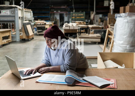 Una femmina nero carpenter lavorando su un computer lap top dopo le ore di lavoro in una grande fabbrica per la lavorazione del legno. Foto Stock