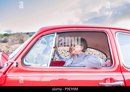 Coppia di anziani con cappello, con occhiali, con il grigio e il bianco dei capelli, con camicia casual, su vintage auto rossa in vacanza godendosi il tempo e la vita. Con un chee Foto Stock