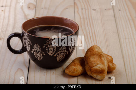 Tazza marrone con ornamenti riempito con tè e due croissant su una luce sullo sfondo di legno. mattina concetto, prima colazione Foto Stock