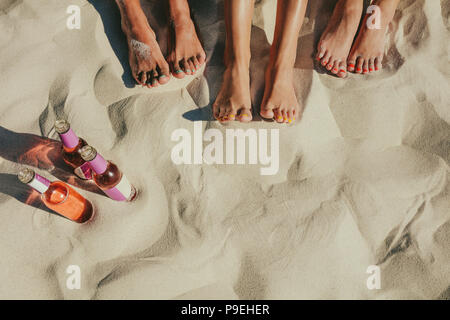 Immagine ravvicinata di gambe di tre donne seduto sulla spiaggia di sabbia con chiodi dipinte in colori diversi e tre bottiglie di bevande analcoliche al loro fianco. Foto Stock