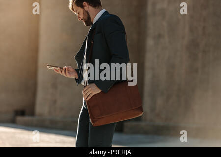 Imprenditore guardando al suo telefono cellulare mentre si cammina sulla strada per l'ufficio. Ufficio occupato andando uomo il pendolarismo a office portando la sua borsa da ufficio e utilizzo Foto Stock