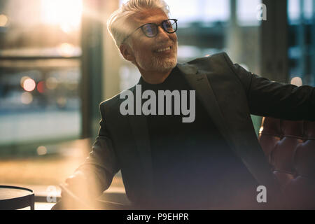 Sorridente imprenditore maturo seduto in ufficio lobby con luce brillante in background. Felice l'uomo senior in business vestiti in seduta office lounge. Foto Stock