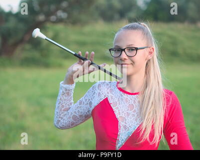 Bespectacled Blonde Teen Majorette ragazza fa roteare Baton all aperto in abito rosso Foto Stock