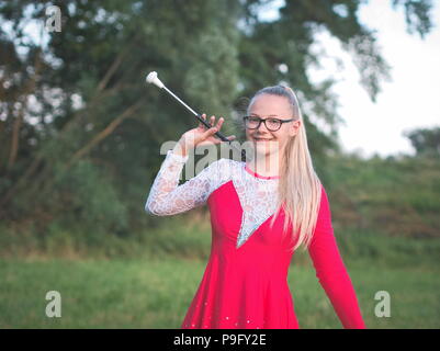 Bespectacled Blonde Teen Majorette ragazza fa roteare Baton all aperto in abito rosso Foto Stock