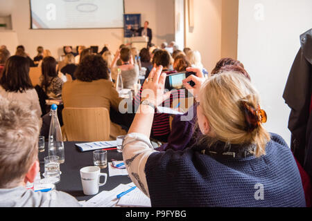 Bianca bionda donna di mezza età può contenere fino il suo smartphone per scattare una foto di una presentazione di Powerpoint in occasione di una conferenza con un sacco di persone in camera Foto Stock