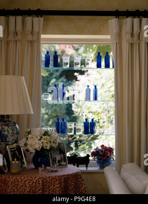 La crema rosa pizzico plissettate oscuranti sulla finestra con una collezione di blu di bottiglie di vetro su ripiani in vetro Foto Stock
