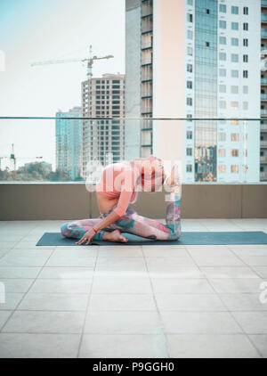 Giovane donna slim con rosa capelli tinti facendo yoga pratica sulla terrazza della città moderna. Ragazza per tenersi in forma e corpo sano relax sulla terrazza sul tetto durante la pratica, rappresentano Foto Stock