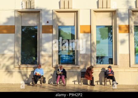 Quattro persone senior maschio e femmina sitrting in serata sole sui sedili di legno posto al di fuori di un grande edificio Faro PORTOGALLO Foto Stock