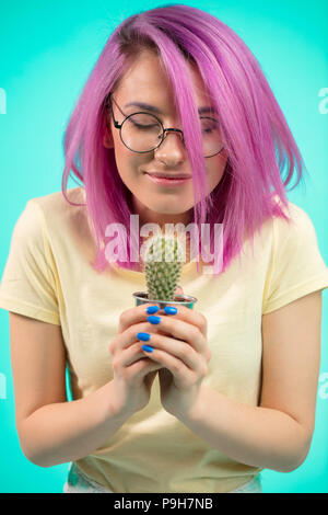 Emotiva dai capelli tinti donna con capelli viola azienda pianta in vaso in mani. Ragazza sorridente indossa occhiali, guardando il cactus con felice allegro expr Foto Stock