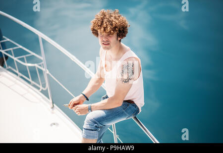 Alta angolazione del bello hipster uomo con spessore capelli ricci, seduti su raillings di una barca a vela, in attesa che i suoi amici di arrivare a una gita in mare ov Foto Stock