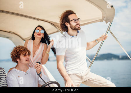 Bello caucasica uomo ricci navigazione di una barca a vela seduto al volante di agitazione, felice elegantemente vestito giovani uomini e donne correre lungo il wa Foto Stock