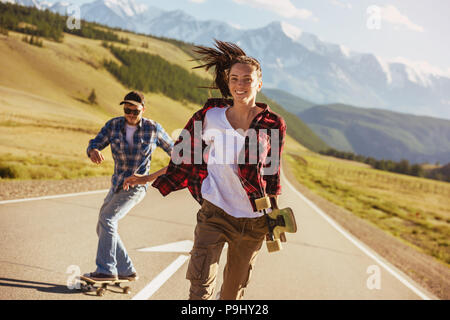 Happy amici si divertono con pattini e longboards a dritta strada contro le montagne Foto Stock