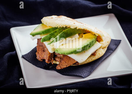 Arepa fatta con avocado, la carne e il formaggio su una piastra bianca e uno sfondo scuro Foto Stock