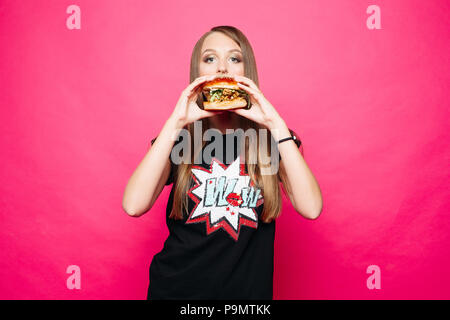 Ritratto in studio di bionda ragazza in black t-shirt con stampa di mangiare hamburger e guardando la telecamera contro vivid magenta sfondo. Foto Stock