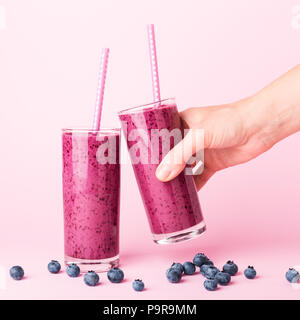 Due bicchieri di mirtilli smoothie con cannucce su sfondo rosa. Donna di mano che tiene un bicchiere. Una sana bevanda estiva. Foto Stock