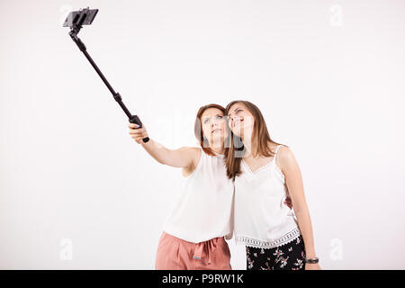 Due bella giovane donna prendendo un selfie con un bastone selfie e produrre diverse divertenti e sciocco volti mentre facendo. Studio foto su un muro bianco Foto Stock