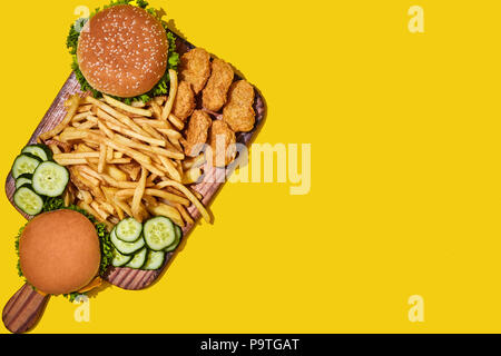Tavola di legno con diversi fast food: hamburger, pepite di pollo e patatine fritte su sfondo giallo. Vista da sopra con copia spazio. Pattern. Lay piatto. Foto Stock