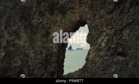 Barca a vela vista attraverso un foro nella roccia. Nave a vela yacht con vele blu nell'oceano. Barca a vela sul mare. Filippine, Boracay. Concetto di viaggio. Foto Stock