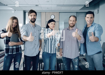 Godetevi ogni giorno. Il gruppo di allegro giovani colleghi thumbing fino e di stare in ufficio Foto Stock