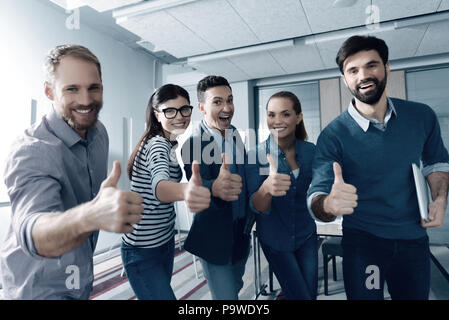 Solo emozioni positive. Allegro sorridente giovani colleghi in piedi in ufficio e thumbing mentre si sentono felici di lavorare insieme Foto Stock