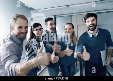 Rimanere ottimista. Positivi i colleghi sorridente sorridente e in piedi in ufficio mentre thumbing fino Foto Stock