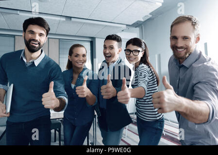 Luogo il vostro umore. Positivo il piacere giovane gruppo di persone thumbing fino e sorridenti mentre in piedi in ufficio Foto Stock