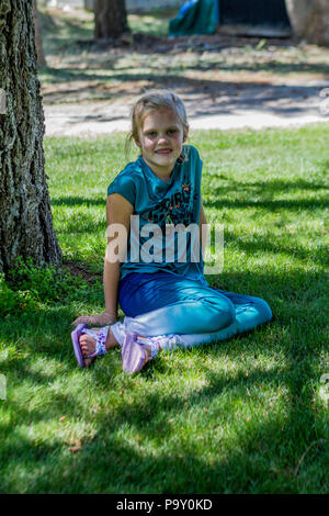 Bella ragazza bionda, in baseball hat e vestito blu, smiljng alla telecamera come si siede nell'ombra di un albero. Modello di Rilascio #113 Foto Stock