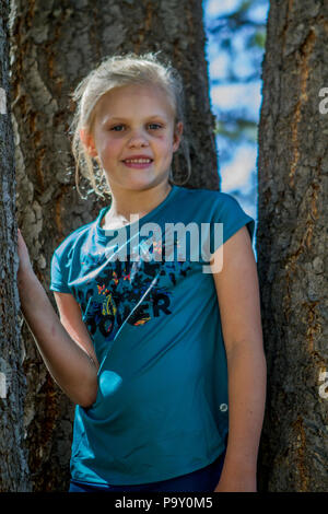 Bella ragazza bionda, in baseball hat e vestito blu, smiljng alla telecamera come si siede nell'ombra di un albero. Modello di Rilascio #113 Foto Stock