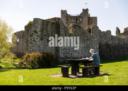 Trim, Irlanda - Uomo seduto su una panchina di picnic e lettura carta di mattina di fronte ad un anglo-normanna Trim Castle, nella contea di Meath. Foto Stock