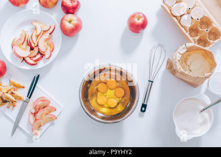 I componenti e gli utensili per la cottura della torta di mele, vista dall'alto. Recipiente metallico riempito con materie tuorli ed albumi vicino a frusta, tagliare la frutta, gusci d'uovo, farina e s Foto Stock