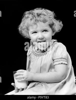 1920s 1930 sorridente bambina ricci capelli corti tenendo un bicchiere di latte guardando la telecamera leccare le sue labbra con linguetta deliziosa - f524 HAR001 ESPRESSIONI HARS B&W contatto visivo leccare la felicità bevanda forza CURLY nutrimento la crescita novellame gustoso in bianco e nero di etnia caucasica deliziosa HAR001 in vecchio stile Foto Stock