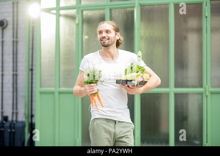 Uomo che porta scatola piena di fresche verdure crude appena comprato al mercato locale all'aperto su sfondo verde Foto Stock