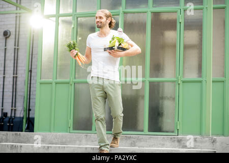 Uomo che porta scatola piena di fresche verdure crude appena comprato al mercato locale all'aperto su sfondo verde Foto Stock