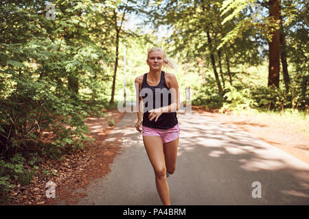 Montare la giovane donna bionda in sportswear focalizzata sulla sua corri da solo su un sentiero di bosco in una giornata di sole Foto Stock