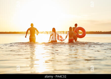 Specchietto di un gruppo di diversi giovani amici in costume da bagno correndo insieme in un lago al tramonto Foto Stock