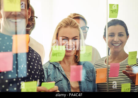 Gruppo sorridente di diversi giovani imprenditori in piedi in un ufficio moderno il brainstorming con foglietti adesivi su una parete di vetro Foto Stock